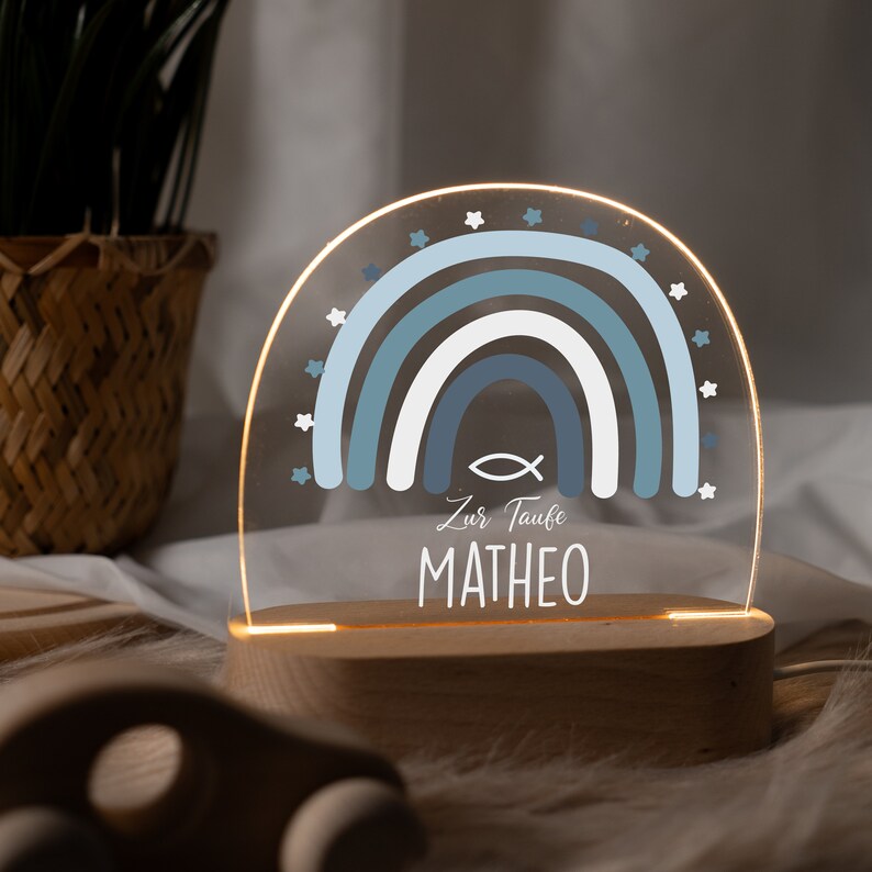 May include: A blue and white rainbow with stars around it, a fish symbol, and the text "Zur Taufe MATHEO" on a wooden base. This is a light-up nightlight.