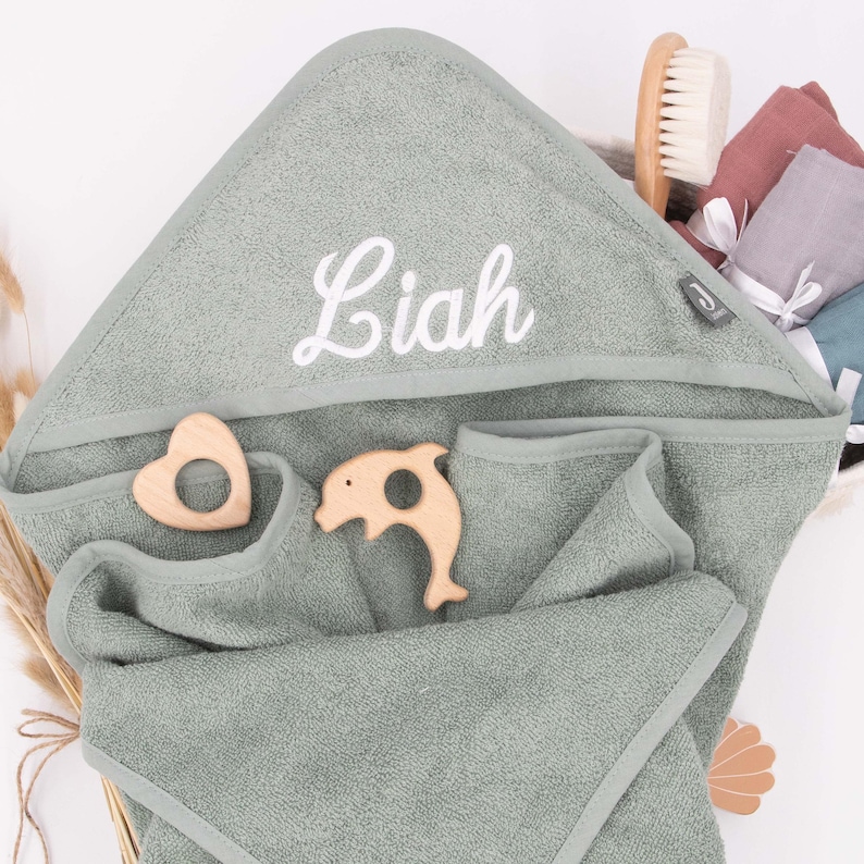 K&ouml;nnte beinhalten: Ein kapuzenhandtuch f&uuml;r Babys in Salbeigr&uuml;n mit dem Namen "Liah" in Wei&szlig; gestickt. Zwei Holzspielzeuge, ein Herz und ein Delfin, sind auf dem Handtuch platziert. Eine Holzb&uuml;rste und andere Babyartikel befinden sich im Hintergrund.
