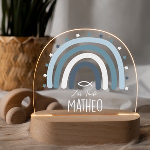 May include: A blue and white rainbow with a fish symbol and the text "Zur Taufe MATHEO" on a wooden base. The rainbow is illuminated from behind, creating a warm glow.