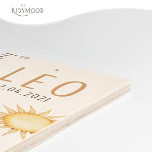 May include: A light-colored wooden growth chart featuring the name LEO in brown letters, the date 07.04.2021, a sun illustration, and measurement markings starting at 150. The brand name KIDSMOOD is printed in the upper left corner.