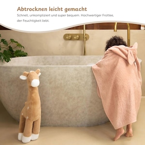 K&ouml;nnte beinhalten: Ein Pl&uuml;sch-Giraffen-Spielzeug und ein rosa Kapuzenhandtuch mit Hasenohren befinden sich in der N&auml;he einer Badewanne. Das Handtuch ist um eine Person gewickelt. Der Text auf dem Bild lautet "Abtrocknen leicht gemacht".