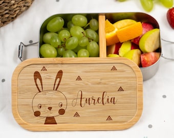 Peut inclure: Une boîte à lunch en bois avec un intérieur en acier inoxydable. Le couvercle est gravé d'une illustration de lapin mignon et du nom "Aurelia". La boîte à lunch est remplie de raisins, de pommes et d'oranges.