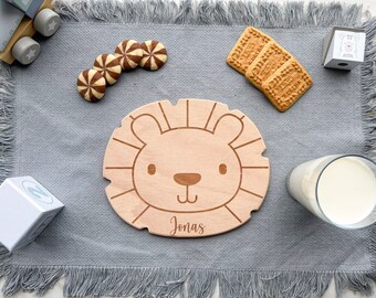May include: A wooden lion-shaped plate with a personalized name "Jonas" engraved on it. The plate is on a gray placemat with a fringe border. There are cookies and a glass of milk on the placemat.