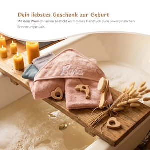 K&ouml;nnte beinhalten: Ein h&ouml;lzernes Badetablett h&auml;lt einen Stapel rosa Kapuzenhandt&uuml;cher mit dem Namen "Cara", zusammen mit einem Holzdelphin, einem herzf&ouml;rmigen Bei&szlig;ring und einer B&uuml;rste. Das Tablett enth&auml;lt auch Trockenblumen und brennende Kerzen.