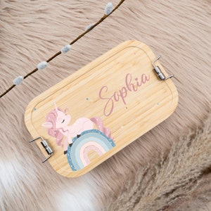 May include: A light brown wooden lunchbox with a pink unicorn and rainbow design. The lunchbox has a metal clasp and the name "Sophia" is written on the front.