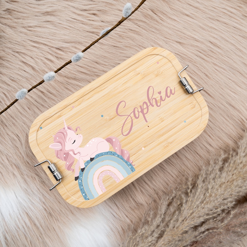 May include: A light brown wooden lunchbox with a pink unicorn and rainbow design. The lunchbox has a metal clasp and the name "Sophia" is written on the front.