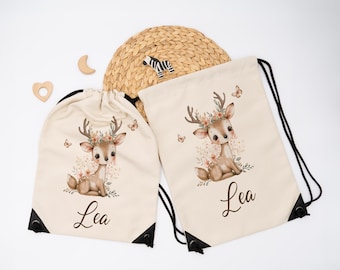 Op de afbeelding: Twee beige trekkoordtassen met een hertenillustratie. Het hert heeft een bloemenkrans en is omringd door bloemen en vlinders. De naam "Lea" staat in script. Zwarte trekkoorden en hoeken.