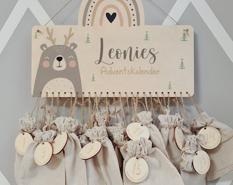 May include: Wooden Advent calendar with a deer and rainbow design. The calendar has 24 numbered pouches for gifts. The name Leonies Adventkalender is written on the top.