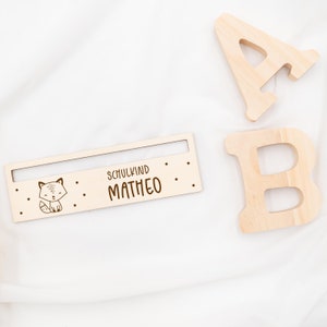 May include: Wooden sign with a fox illustration and the text "Schulkind Matheo". The sign is light brown and has a slot for a name card.