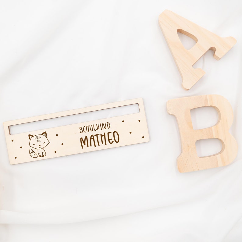 May include: Wooden sign with a fox illustration and the text "Schulkind Matheo". The sign is light brown and has a slot for a name card.