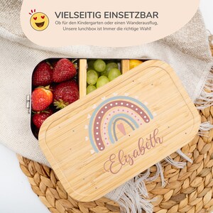 Może przedstawiać: Drewniana lunchbox z wzorem tęczy i imieniem "Elisabeth" na pokrywce. Lunchbox jest otwarta i wypełniona truskawkami i winogronami.