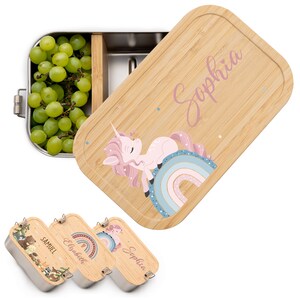 May include: A set of three stainless steel lunchboxes with bamboo lids. Each lid features a different design: a unicorn on a rainbow, a forest scene, and a rainbow with stars. The lids are personalized with the names "Sophia", "Elisabeth", and "Samuel".