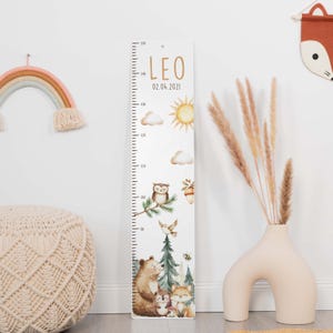 May include: A vertical wooden growth chart featuring the name LEO and the date 02.04.2021. The ruler displays measurements from 90 to 150 cm, decorated with illustrations of a sun, clouds, an owl, a bear, a hedgehog, a fox, and pine trees.