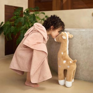 K&ouml;nnte beinhalten: Ein Kind in einem hellrosa Kapuzenhandtuch k&uuml;sst eine Stoffgiraffe. Die Giraffe ist hellbraun mit wei&szlig;en Flecken und Akzenten. Der Hintergrund ist neutral. Das Handtuch ist aus weichem Material.