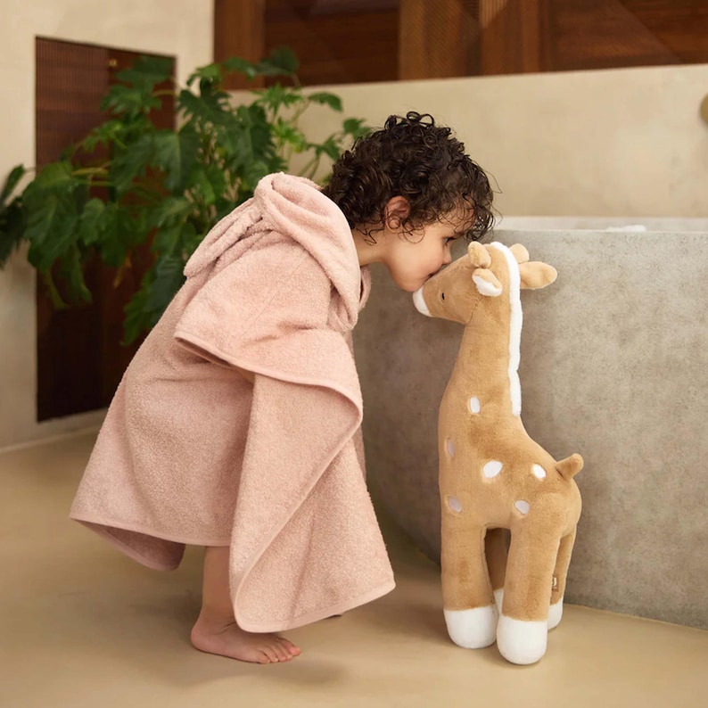 K&ouml;nnte beinhalten: Ein Kind in einem hellrosa Kapuzenhandtuch k&uuml;sst eine Stoffgiraffe. Die Giraffe ist hellbraun mit wei&szlig;en Flecken und Akzenten. Der Hintergrund ist neutral. Das Handtuch ist aus weichem Material.