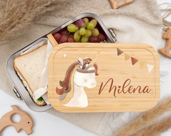 Peut inclure: Une boîte à lunch en acier inoxydable avec un couvercle en bambou orné d'un motif de licorne à l'aquarelle et du nom "Milena". La boîte à lunch contient un sandwich et des raisins. Le couvercle présente un motif de bannière marron et beige.