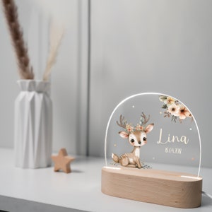 Op de afbeelding: Een houten nachtlampje met een heldere acryl inzet met een cartoon hert met een bloemenkrans. Het hert is bruin met witte vlekken en zit. De acryl inzet heeft ook een bloemenontwerp en de naam "Lina" met de datum "18.04.2018" eronder.