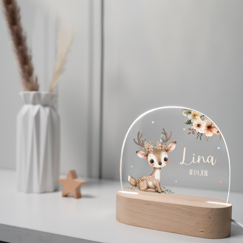 Op de afbeelding: Een houten nachtlampje met een heldere acryl inzet met een cartoon hert met een bloemenkrans. Het hert is bruin met witte vlekken en zit. De acryl inzet heeft ook een bloemenontwerp en de naam "Lina" met de datum "18.04.2018" eronder.