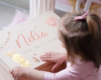 May include: A wooden keepsake box with the name "Nelia" in pink script. The box is decorated with a bunny, hot air balloon, and a banner. The box also includes the date, weight, and length of the baby.