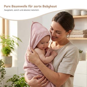 K&ouml;nnte beinhalten: Ein Baby, eingewickelt in ein weiches, rosa Kapuzenhandtuch, wird von einer Person gehalten. Das Handtuch besteht aus saugf&auml;higem Material. Der Text oben im Bild lautet: "Reine Baumwolle f&uuml;r zarte Babyhaut."