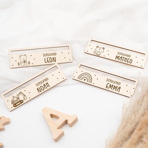 May include: Four wooden nameplates with different designs and names. The names are Leon, Noah, Emma, and Matheo. Each nameplate has a small illustration, such as a bear, an excavator, a rainbow, and a fox.