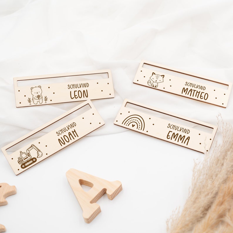 May include: Four wooden nameplates with different designs and names. The names are Leon, Noah, Emma, and Matheo. Each nameplate has a small illustration, such as a bear, an excavator, a rainbow, and a fox.