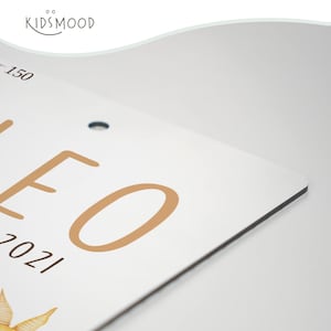 May include: A white rectangular sign with rounded corners featuring the tan text LEO and 2021, along with a partial illustration of a star. The top left corner displays the text KIDSMOOD and the number 150. A small circular hole is visible.