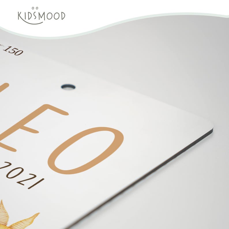 May include: A white rectangular sign with rounded corners featuring the tan text LEO and 2021, along with a partial illustration of a star. The top left corner displays the text KIDSMOOD and the number 150. A small circular hole is visible.