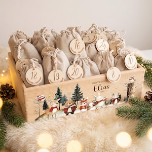 Puede incluir: Una caja de madera que contiene veinticuatro bolsas de tela beige con cordón, cada una con una etiqueta redonda de madera numerada del 1 al 24. La caja presenta el texto "ADVENTSKALENDER Elias" y una ilustración de animales del bosque.