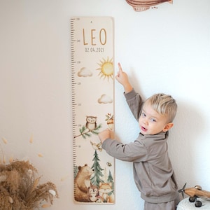 Può includere: Un'asta di crescita in legno a tema bosco, con un orso, una volpe, un gufo e altri elementi della foresta. L'asta è contrassegnata da misure in centimetri e include il nome "LEO" e la data "02.04.2021". Un bambino indica l'asta.