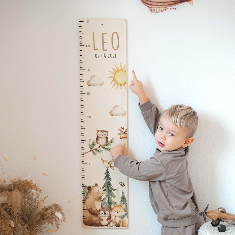 May include: A vertical wooden growth chart featuring a printed scale from 90 to 150 cm, the name LEO, the date 02.04.2021, and illustrations of a sun, clouds, an owl, a bear, a hedgehog, a fox, and pine trees in soft colors.