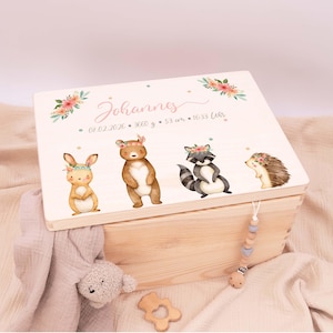 Può includere: Scatola ricordo in legno chiaro con coperchio incernierato. Il coperchio presenta illustrazioni ad acquerello di un coniglietto, un orso, un procione e un riccio, ognuno con una corona floreale. La scatola è personalizzata con un nome e dettagli di nascita.
