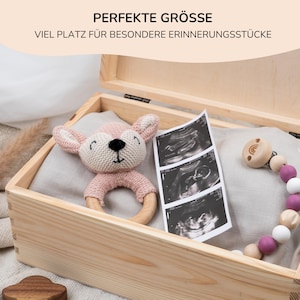 May include: A wooden keepsake box with a light brown finish. The box is open and contains a pink knitted deer rattle, a set of ultrasound images, and a wooden teething toy with pink and white beads.