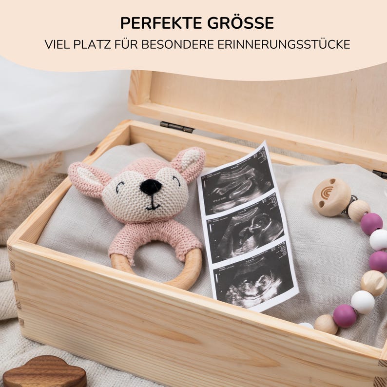 May include: A wooden keepsake box with a light brown finish. The box is open and contains a pink knitted deer rattle, a set of ultrasound images, and a wooden teething toy with pink and white beads.