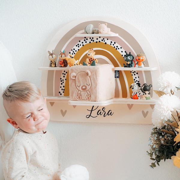 May include: A wooden rainbow shelf with three tiers, decorated with black polka dots and a yellow, orange, and pink ombre design. The shelf is filled with small toys, including a stuffed bear, a stuffed animal, and a small figurine. The shelf is labeled "Lara".