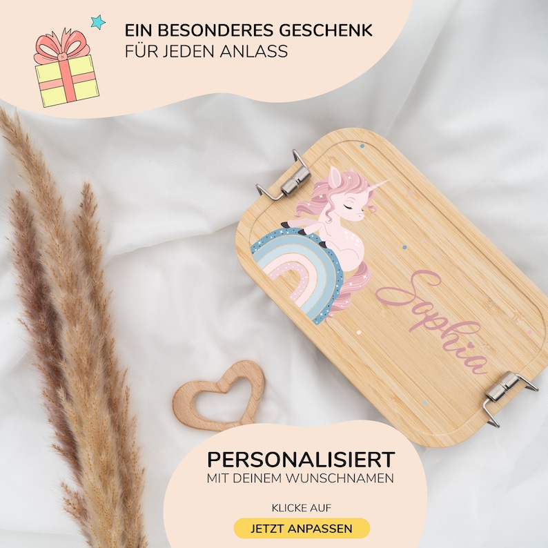 May include: A personalized wooden lunch box with a unicorn and rainbow design. The lunch box is light brown with a pink unicorn and a rainbow with blue, pink, and white colors. The name "Sophia" is written in pink on the lunch box.