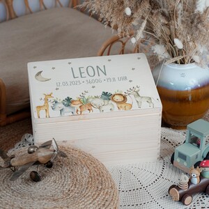 May include: A white wooden keepsake box with a lid featuring a whimsical illustration of animals, including a giraffe, rhino, lion, and zebra. The lid is decorated with stars and a crescent moon. The box is personalized with the name "LEON" and the date "12.03.2023 - 3600G - 19:11 UHR".