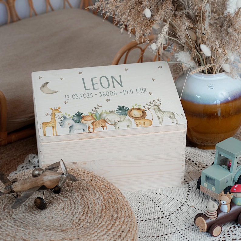 May include: A white wooden keepsake box with a lid featuring a whimsical illustration of animals, including a giraffe, rhino, lion, and zebra. The lid is decorated with stars and a crescent moon. The box is personalized with the name "LEON" and the date "12.03.2023 - 3600G - 19:11 UHR".