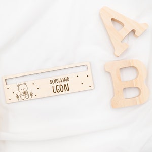 May include: Wooden sign with a bear illustration and the text "Schulkind LEON". The sign is on a white surface with two wooden letters, A and B, in the background.