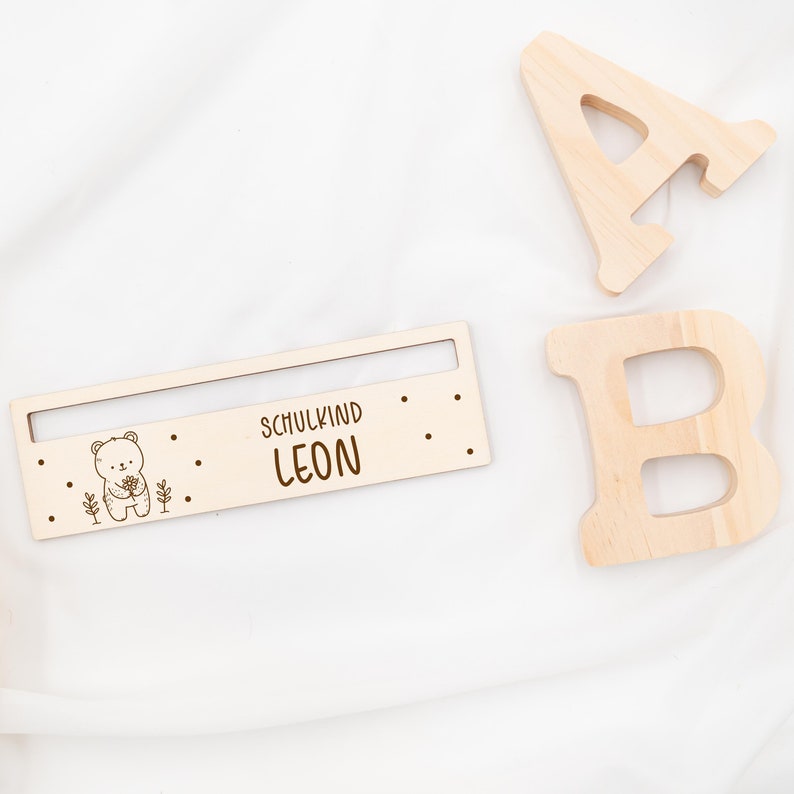 May include: Wooden sign with a bear illustration and the text "Schulkind LEON". The sign is on a white surface with two wooden letters, A and B, in the background.