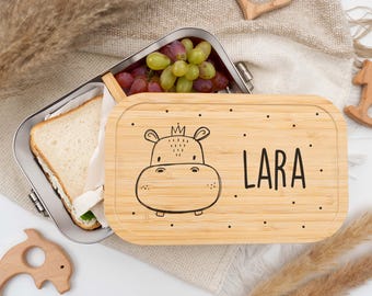 Peut inclure: Une boîte à lunch en bambou avec une illustration d'hippopotame et le nom "LARA" en noir. Le récipient en acier inoxydable contient un sandwich et des raisins. Des jouets en bois en forme de cœur et de dauphin sont également visibles.