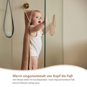 K&ouml;nnte beinhalten: Ein Baby in einem wei&szlig;en Body mit einem braunen Kapuzenhandtuch, das nach einer Holzb&uuml;rste greift. Der Text "Warm eingemummelt von Kopf bis Fu&szlig;" ist unten im Bild zu sehen.