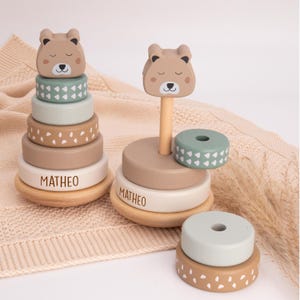 May include: Wooden stacking toy featuring a bear head topper and colorful rings. The rings are in shades of green, beige, and white, with the name "MATHEO" on the base. The toy is designed for young children.