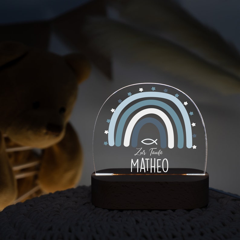 May include: A blue and white rainbow with stars and a fish symbol, a wooden base, and the text "Zur Taufe MATHEO".