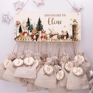 Peut inclure: Un calendrier de l'Avent en bois avec une scène de forêt peinte et le nom "Elias". Vingt-quatre sacs en tissu beige sont suspendus en dessous, chacun avec une étiquette en bois numérotée de 1 à 24. Le calendrier est décoré d'animaux de la forêt et d'étoiles.
