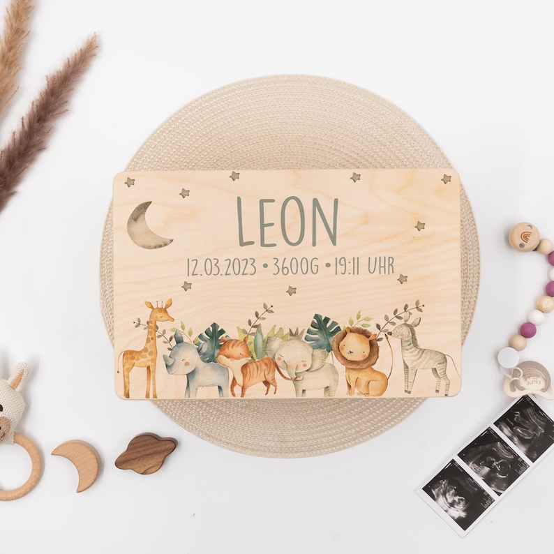 May include: A wooden plaque with a crescent moon cutout and a jungle animal theme. The plaque is personalized with the name "LEON" and the date "12.03.2023", weight "3600g", and time "19:11 UHR".