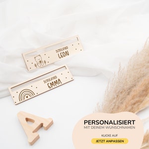 May include: Two personalized wooden nameplates with a bear and rainbow design. The nameplates say "Schulkind Leon" and "Schulkind Emma".