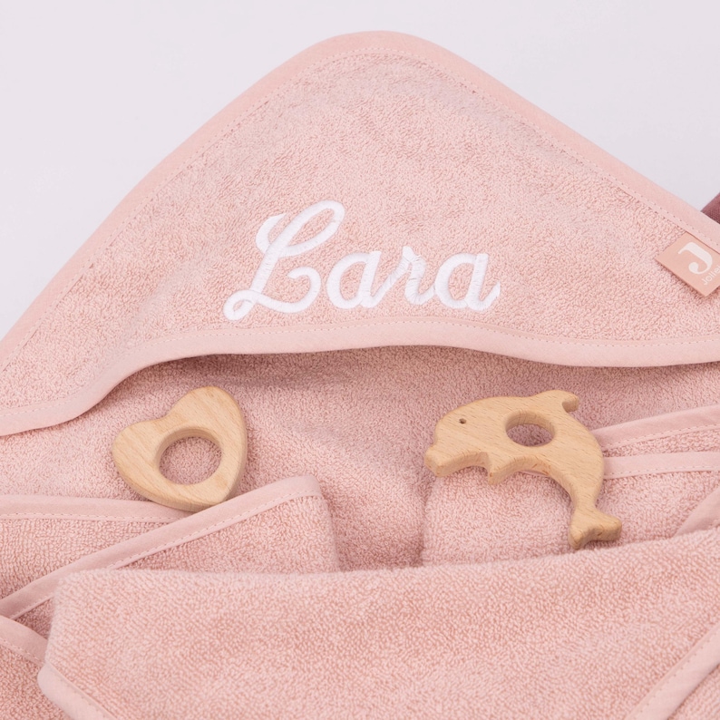 K&ouml;nnte beinhalten: Ein hellrosa Baby-Kapuzenhandtuch mit dem Namen "Lara" in Wei&szlig; gestickt. Zwei Holzspielzeuge, ein Herz und ein Delfin, liegen neben dem Handtuch. Das Handtuch ist aus weichem Frottee.