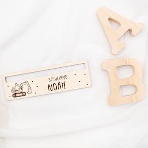 May include: Wooden sign with a cartoon excavator and the text "Schulkind Noah". The sign is light brown and has a small slot at the top.