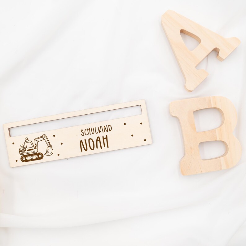 May include: Wooden sign with a cartoon excavator and the text "Schulkind Noah". The sign is light brown and has a small slot at the top.